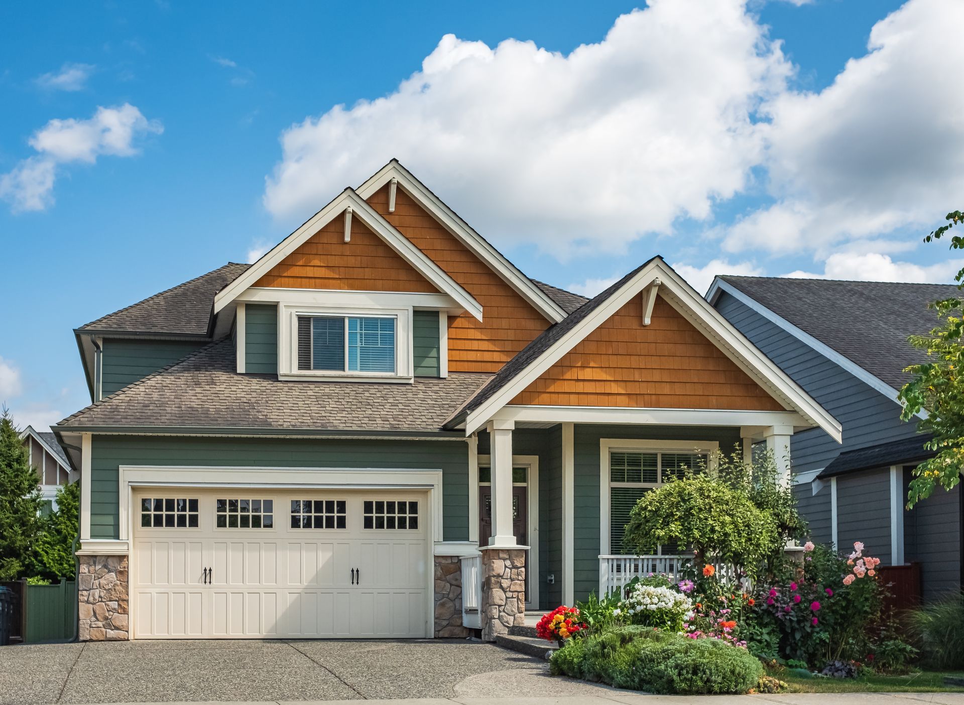 A suburban two-story home with a tan garage, sage green siding, wood-paneled gables, and a lush front garden.
