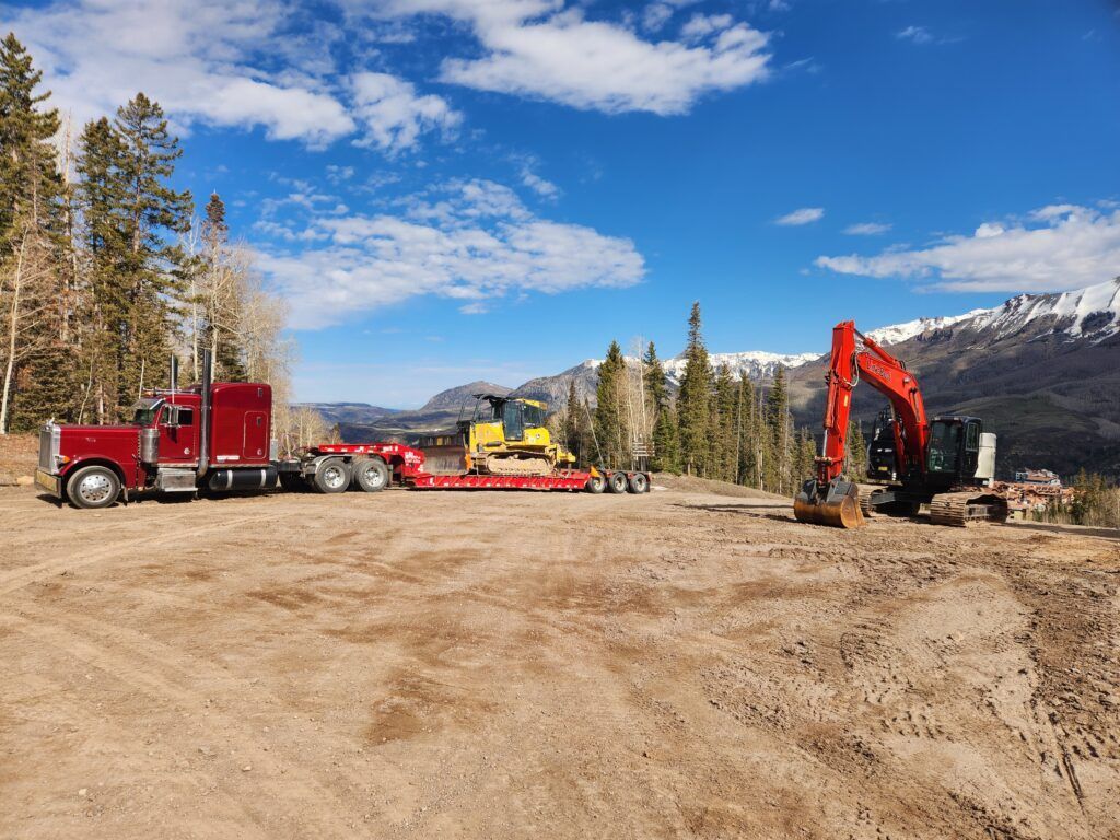 Construction trucks and excavator on a dirt worksite with mountains and pine trees under a blue sky