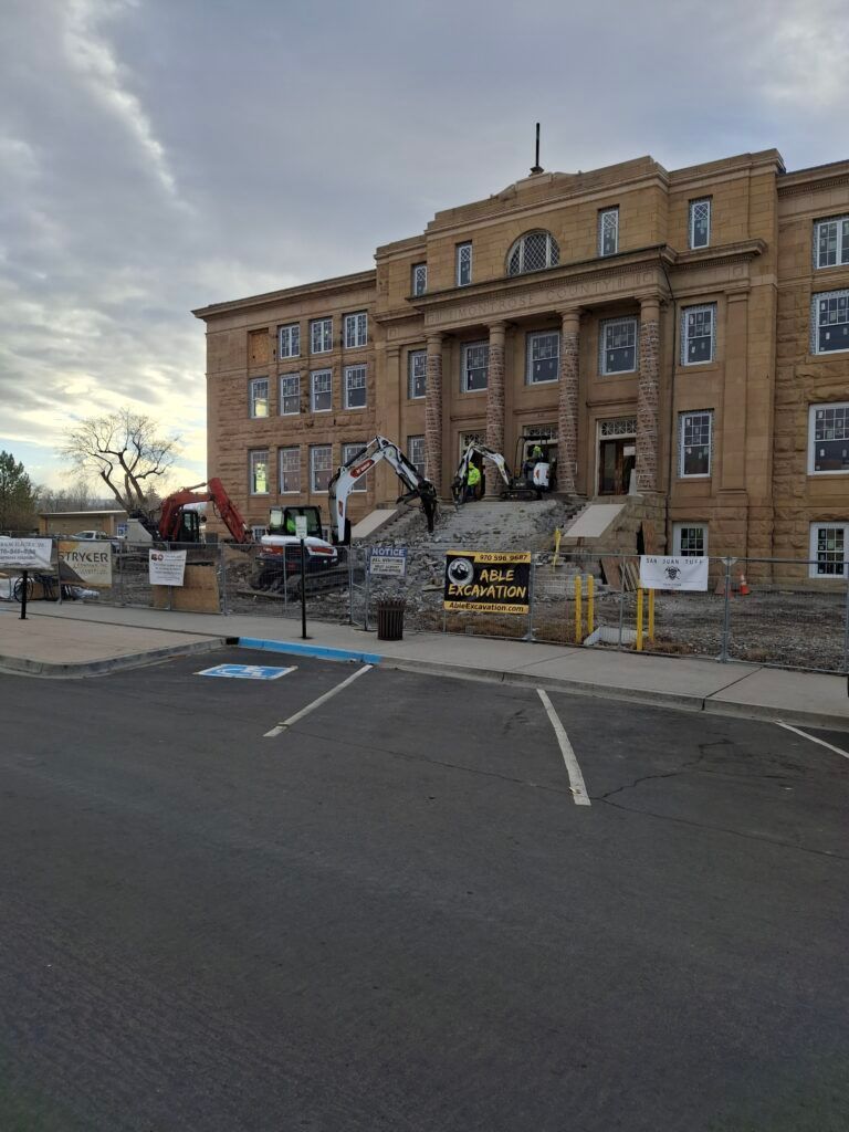 Historic brick building with workers and equipment outside during renovation, viewed from a parking lot.