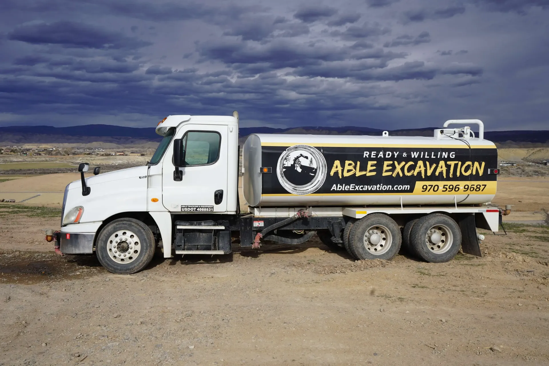 White Able Excavating dump truck parked on a dirt lot under a cloudy sky.