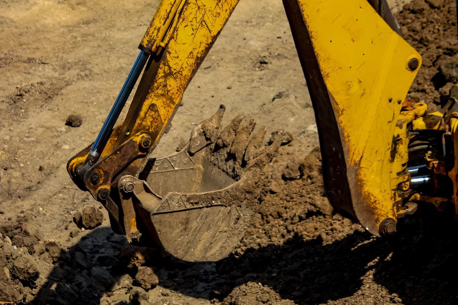 Yellow backhoe bucket digging into dry brown soil while cutting a drainage ditch on a job site