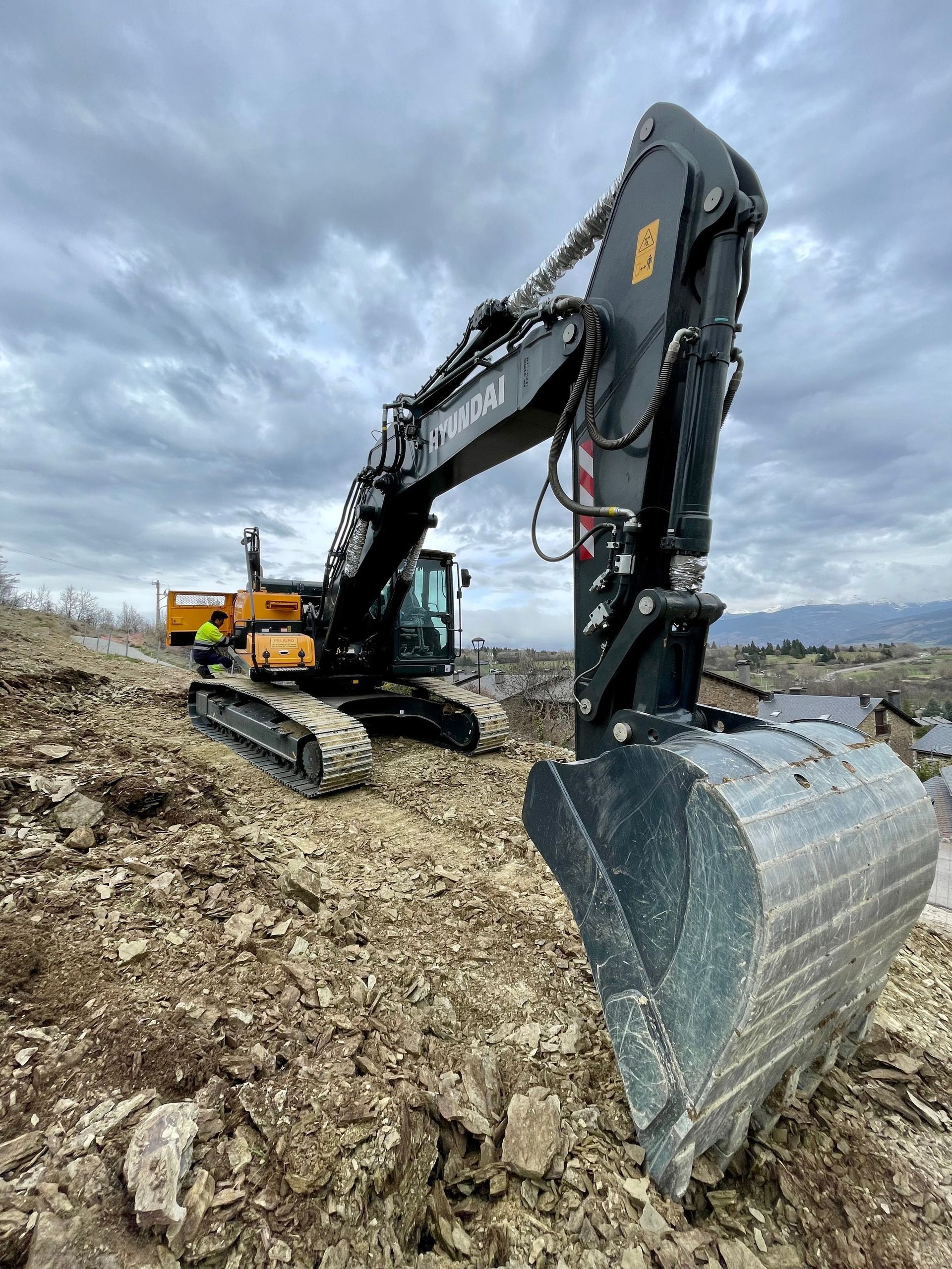 Large excavator on a rocky construction site under a cloudy sky, bucket raised in the foreground.