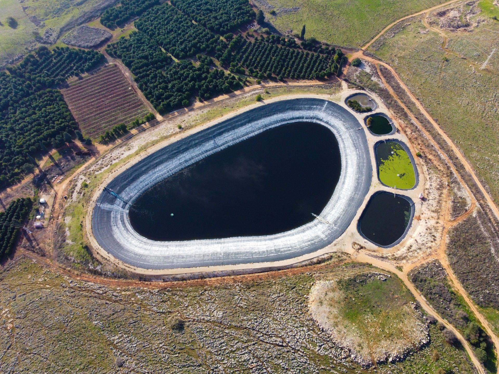 Aerial view of a large oval reservoir surrounded by rocky terrain, fields, and trees.