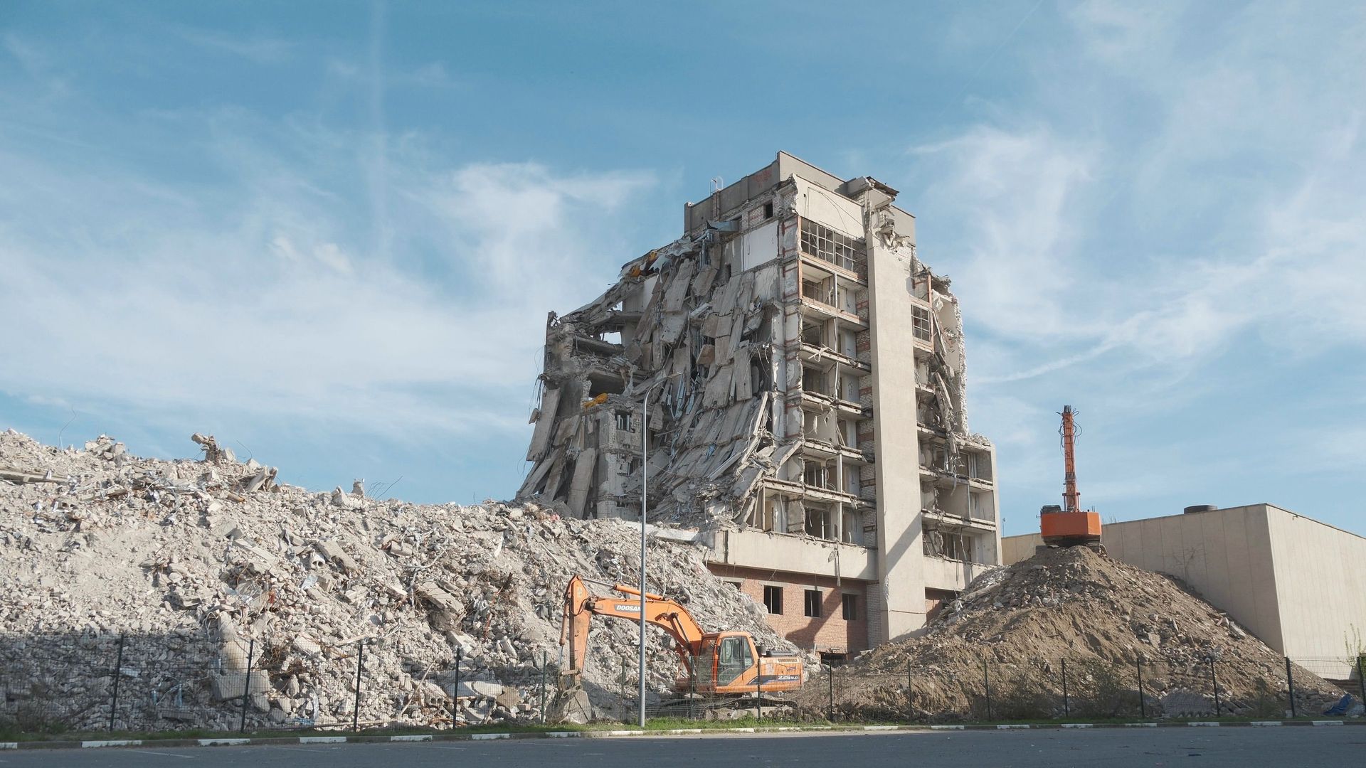 Partially demolished concrete building beside a large pile of rubble under a blue sky