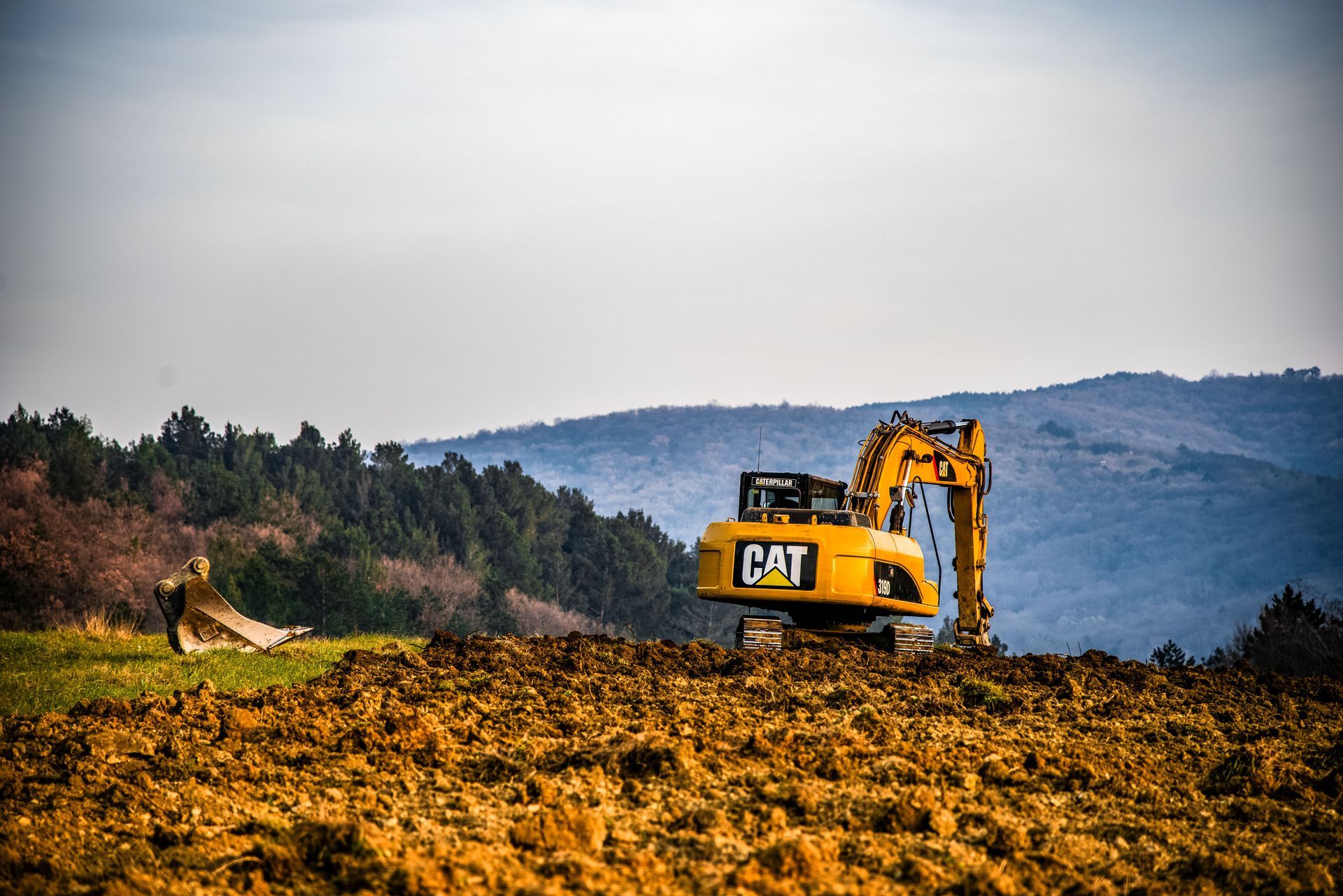 Yellow CAT excavator working in a muddy field with forested hills in the background