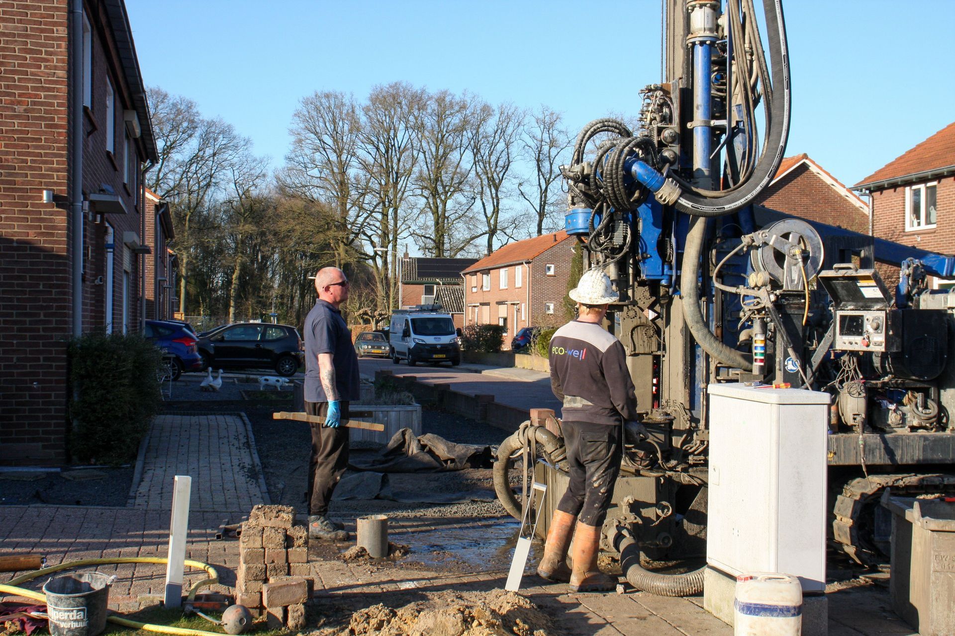 Workers operate a drilling rig on a residential street beside brick houses in sunlight.