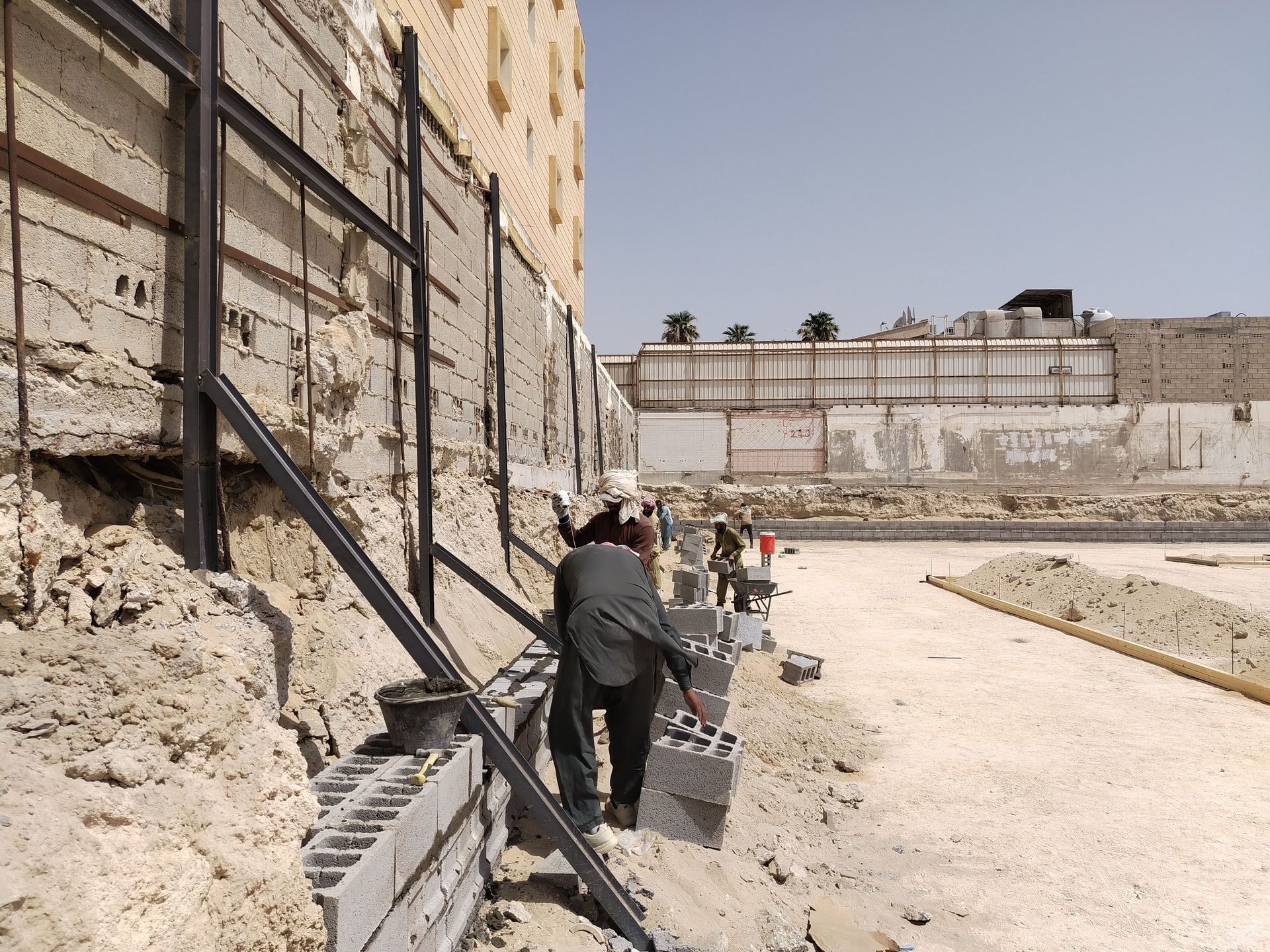 People walking along a dusty path beside a tall retaining wall in a construction site.