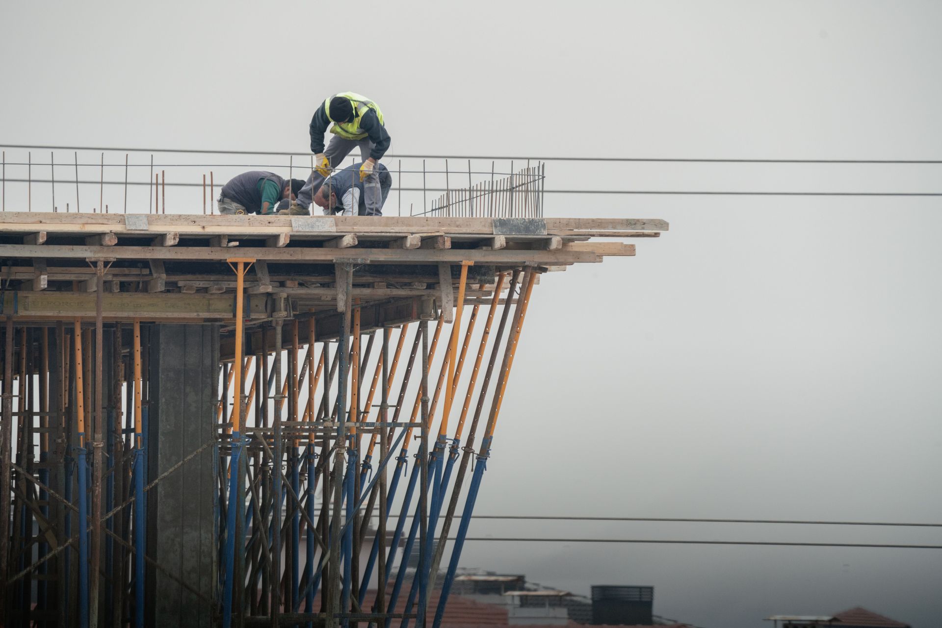 Construction workers on a high-rise platform, bending over the roof edge with scaffolding below