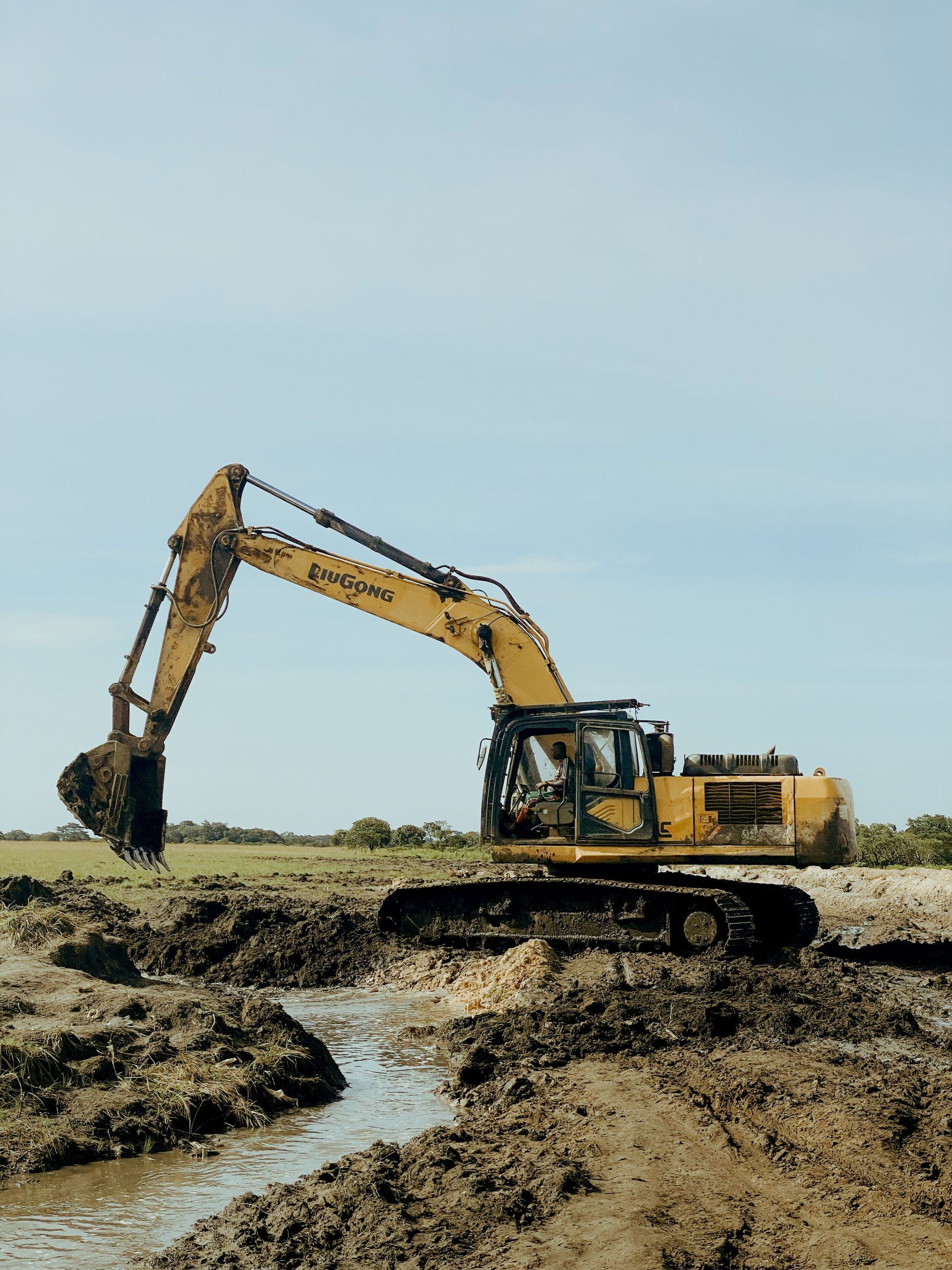 Yellow excavator digging in muddy marshland beside a narrow stream under a cloudy sky