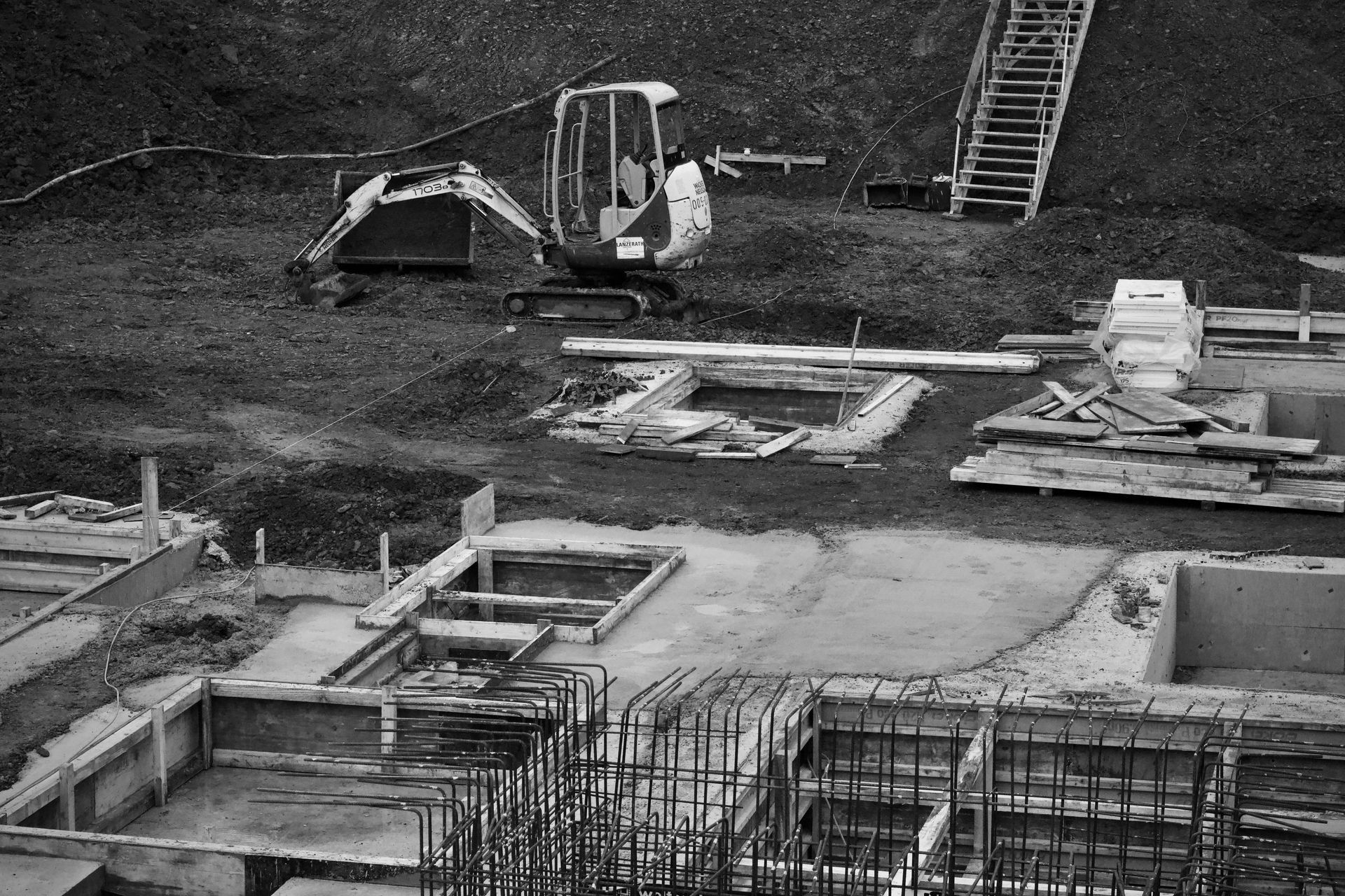 Black-and-white construction site with an excavator, rebar, and concrete foundation forms