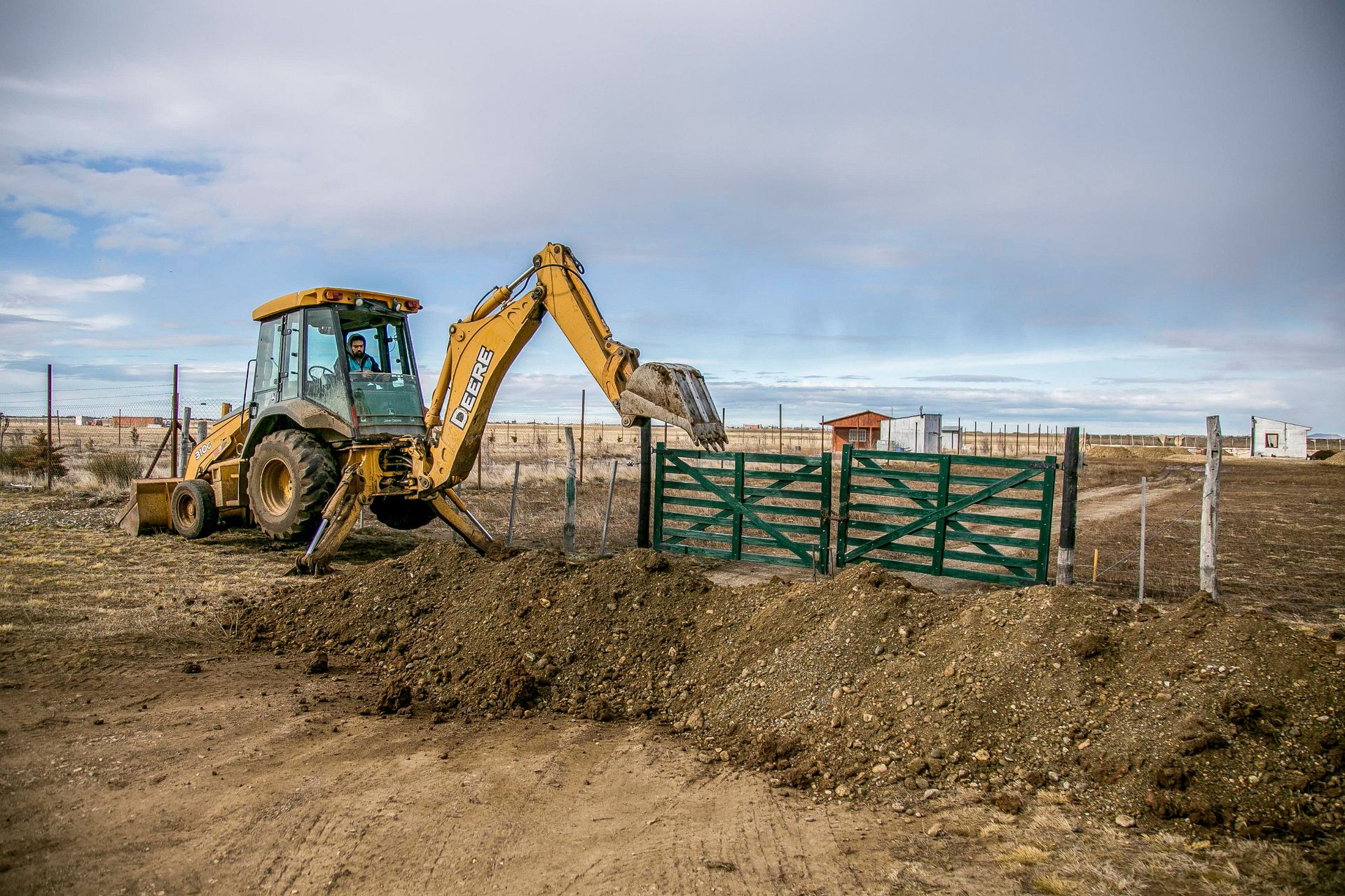 Backhoe and tractor at a fenced dirt construction site under cloudy sky