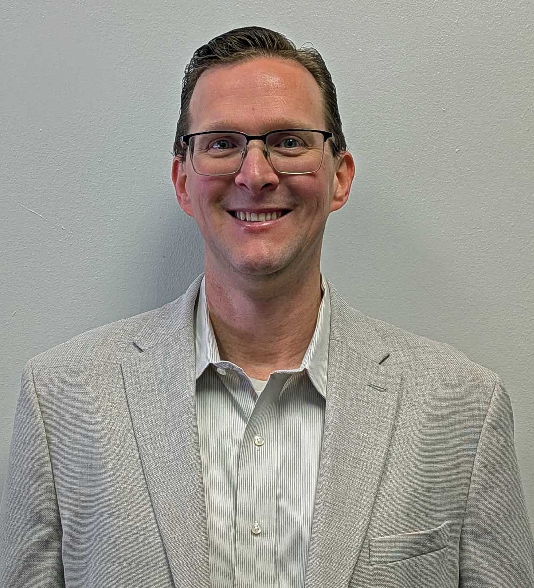 Barrett Rodriguez wearing glasses, light jacket, and button-down shirt smiles in front of a neutral wall.