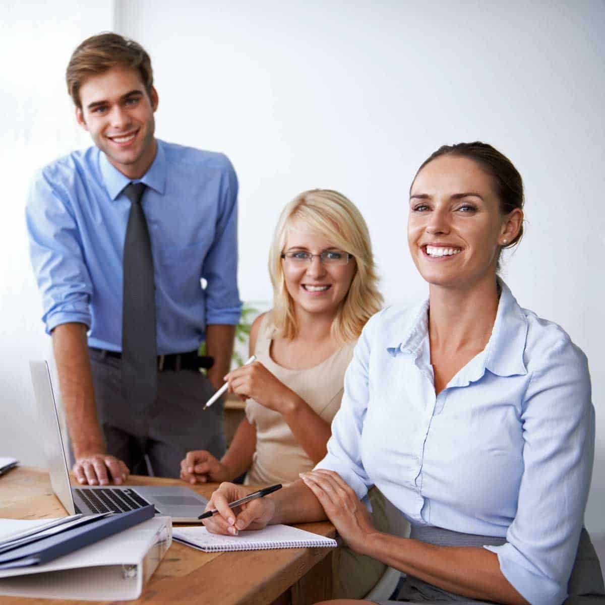 Three smiling coworkers at a desk, looking at the camera.