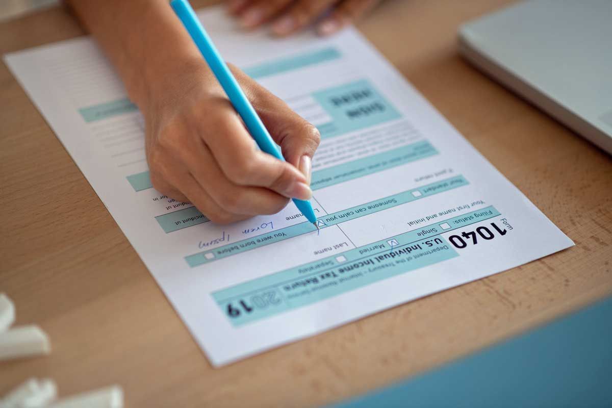 Person's hand with a blue pen filling out a 1040 tax form on a wooden desk.