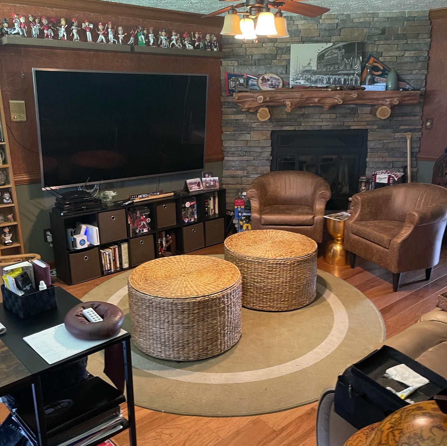 A living room features two tan leather chairs, two round woven ottomans, a large flat-screen TV, and a stone fireplace.