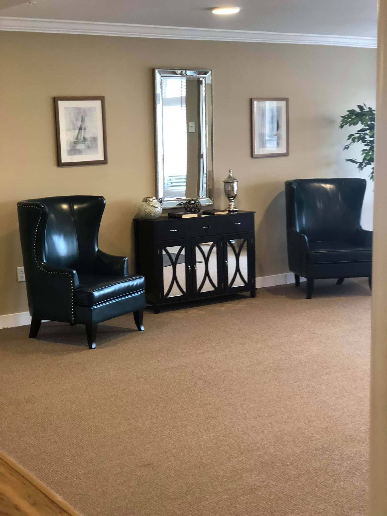 A room featuring a black console table with a mirror between two black leather wingback chairs and two framed prints.