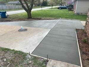 Freshly poured gray concrete walkway beside a driveway and yard, with a small trowel on the slab.