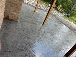 Wet concrete patio under a covered area, with wooden posts and brick walls beside a yard.