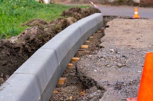 Damaged road curb with cracked pavement and orange traffic cones marking repairs