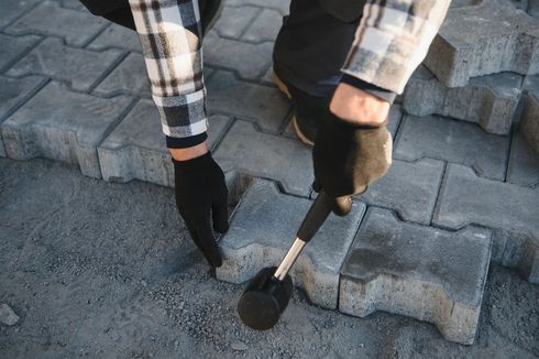 Gloved hands laying gray paving stones on a sidewalk with a mallet nearby