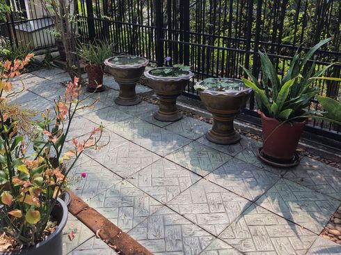 Small patio with patterned stone tiles, four urn planters, and potted tropical plants beside a black fence.