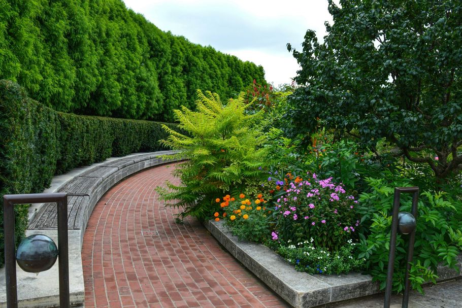 Curved brick garden path beside lush green hedges and colorful flowers, with stone edging and lamps