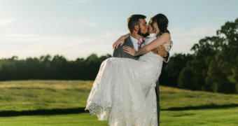 Bride and groom embrace in a grassy field at sunset, with trees in the background.