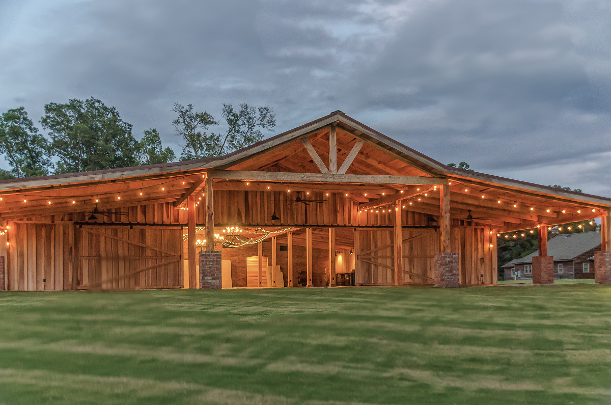 Rustic wooden barn venue lit warmly at dusk, with a wide green lawn and trees behind it.