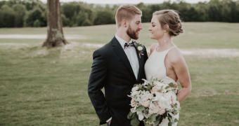 Bride and groom posing outdoors in a grassy field, holding a bouquet and facing each other