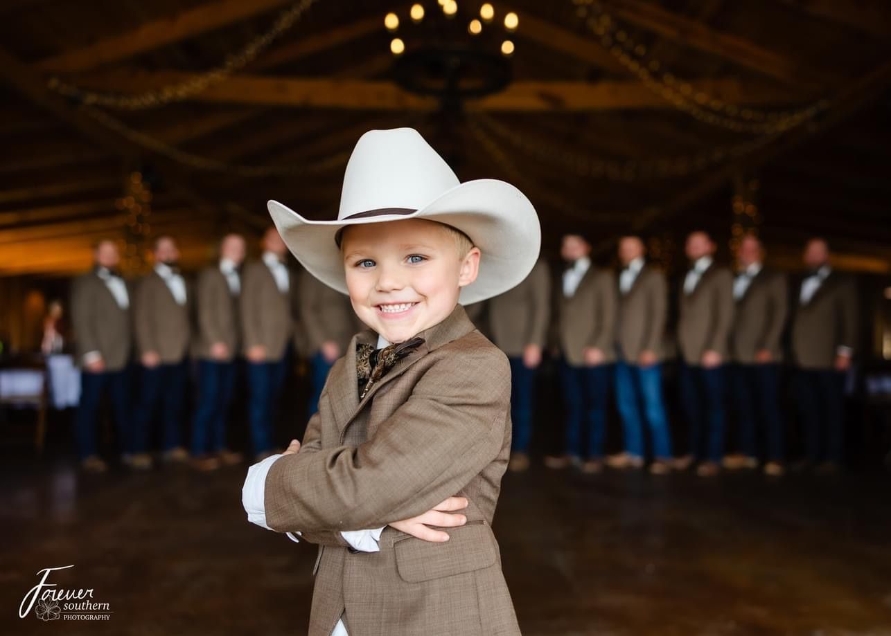 Smiling child in a white cowboy hat and brown suit, standing in a barn with people blurred in the background