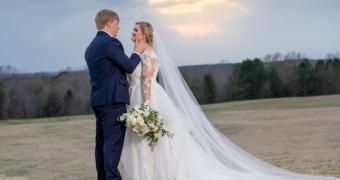 Bride and groom sharing a kiss in a field at sunset, with a long white veil flowing behind her
