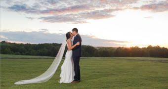 Bride and groom embracing in a grassy field at sunset, with the bride’s veil flowing behind them