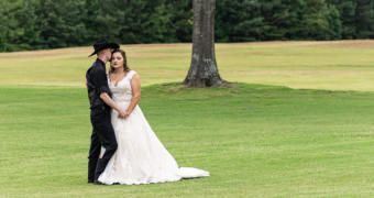 Couple in wedding attire posing on a grassy lawn near a tree