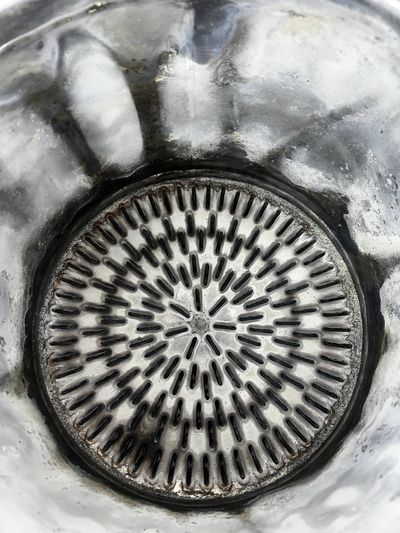 Top-down view of a circular, patterned metal drain grate inside a stainless steel sink.