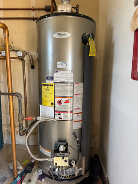 A silver Whirlpool gas water heater with copper plumbing connections standing in a room.