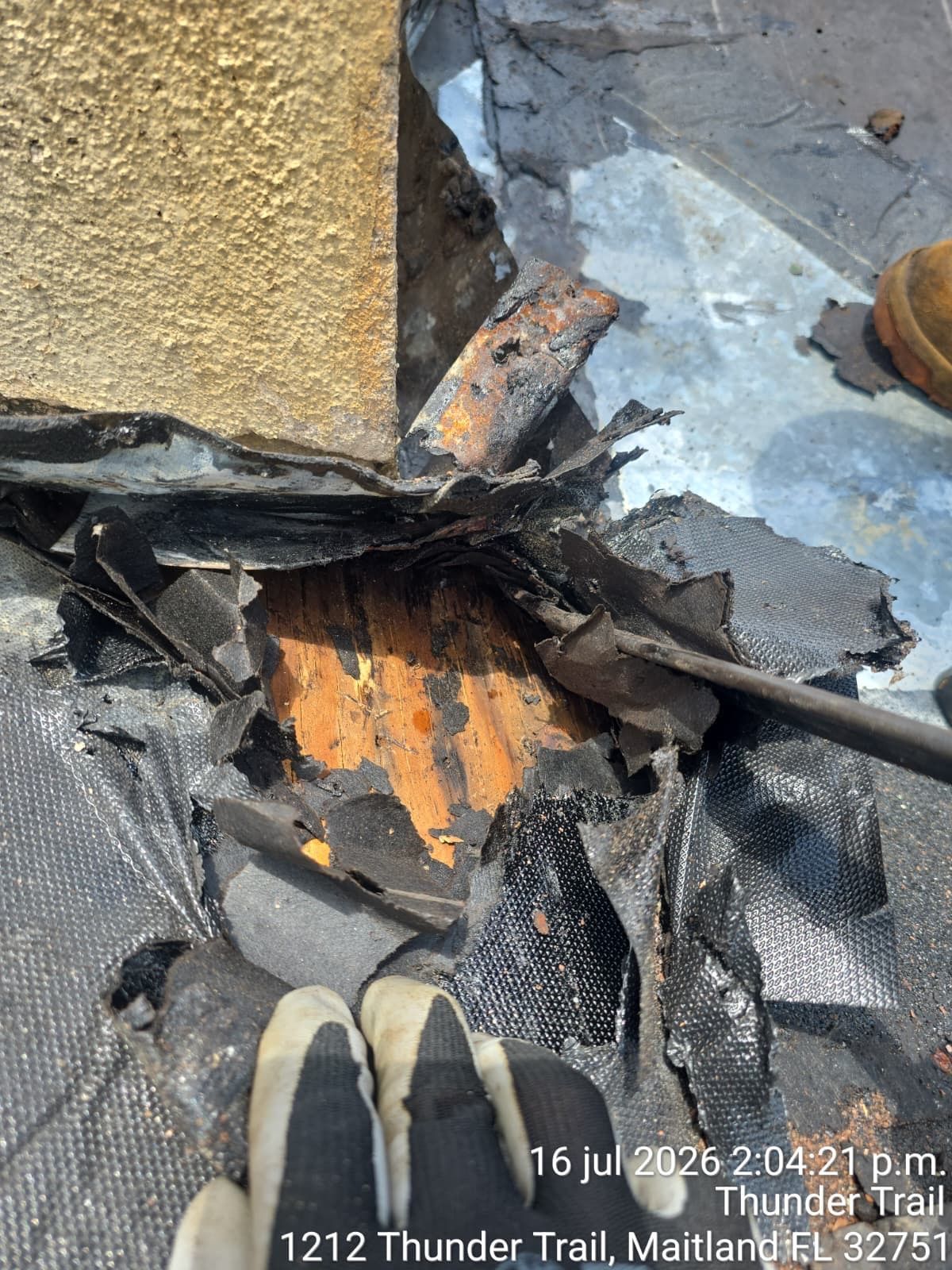 Roof damage with torn black tarp, exposed orange underlayer, and debris beside a wall and pipe.