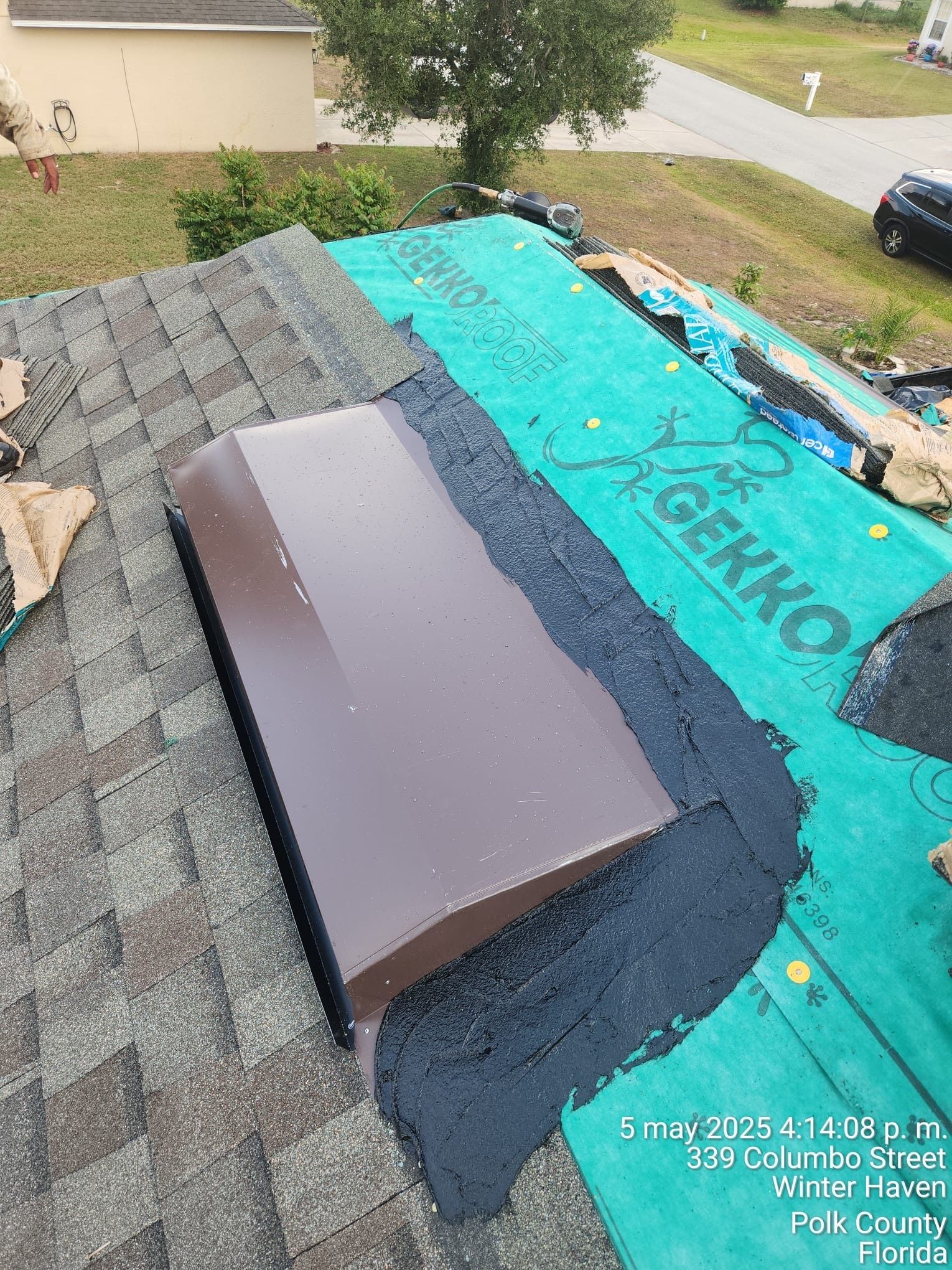 Roof with gray shingles and a vent opening covered by turquoise underlayment near a driveway.