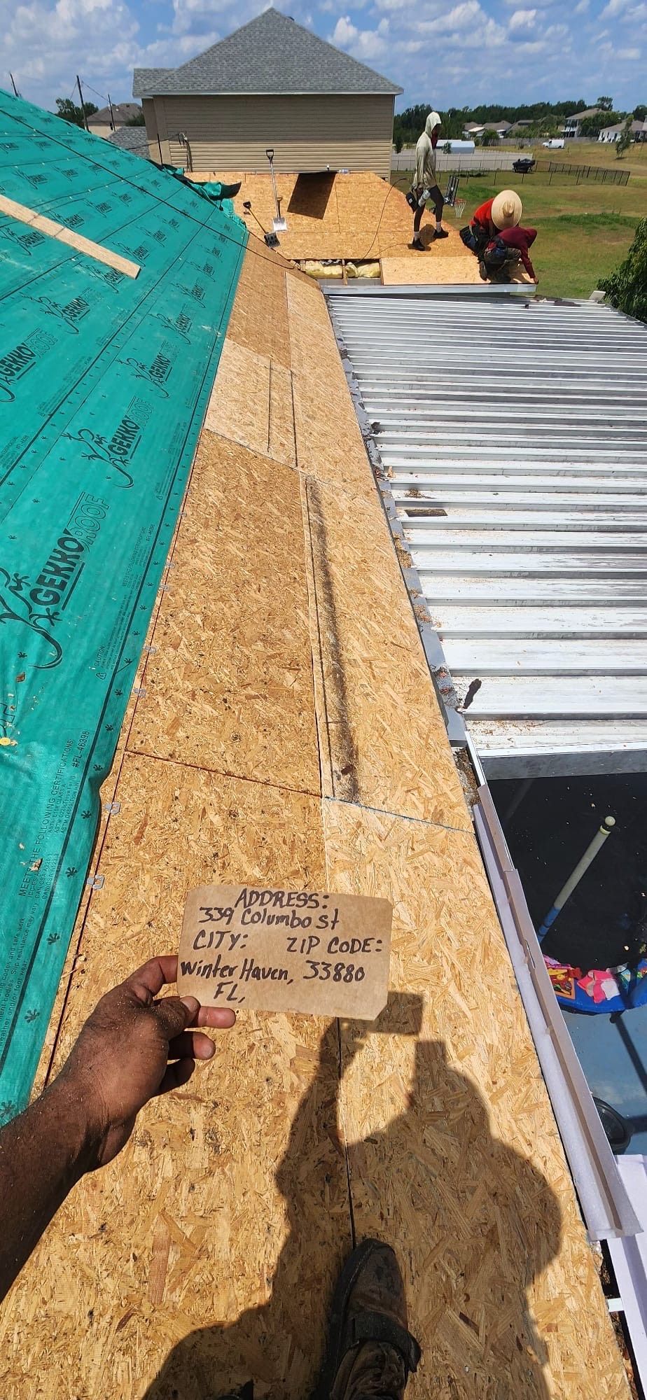 Roofing workers installing panels on a steep roof, with one holding a nailer near the ridge.