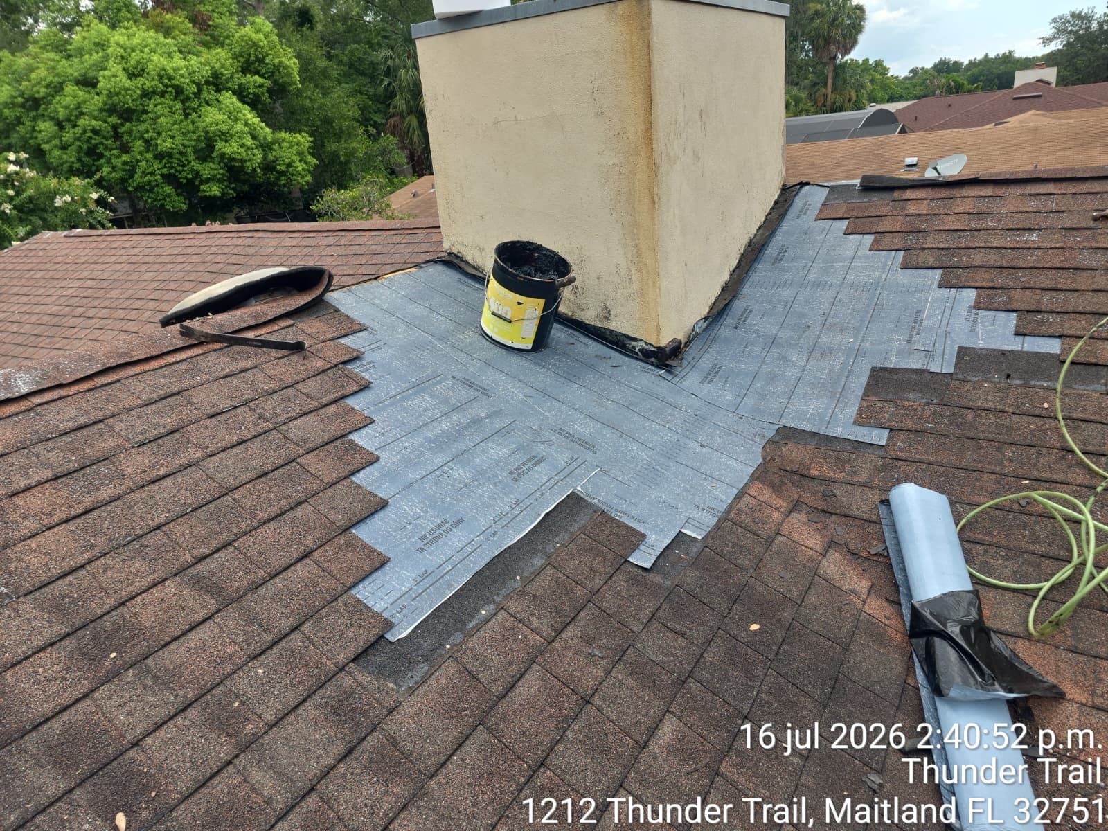 Roof repair around chimney with gray flashing and brown shingles, plus a black bucket and roofing roll.