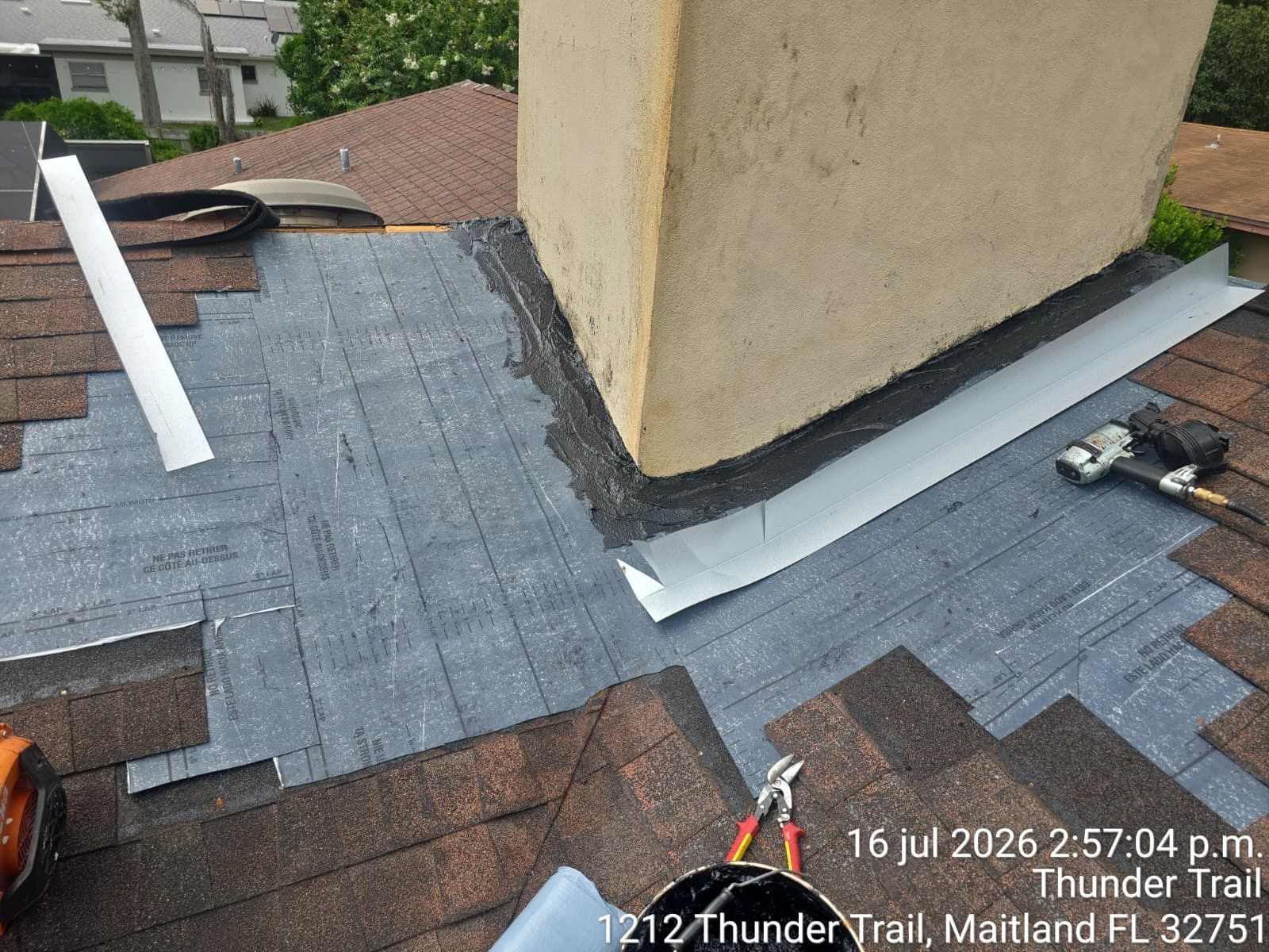 Roofing work around a chimney with gray shingles, white flashing, and tools on a roof deck.