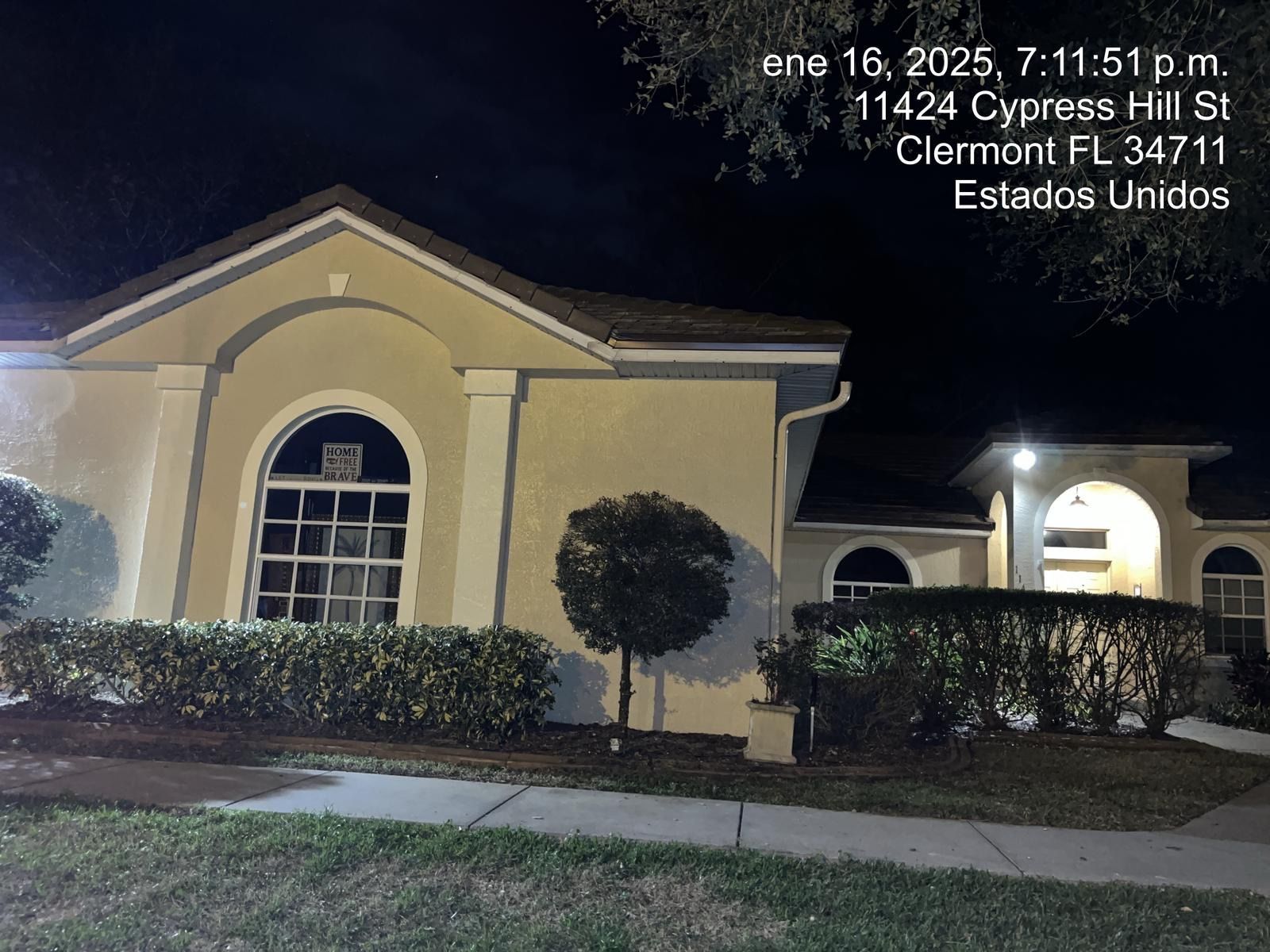 Night view of a beige house with lit windows, manicured shrubs, and a sidewalk in front