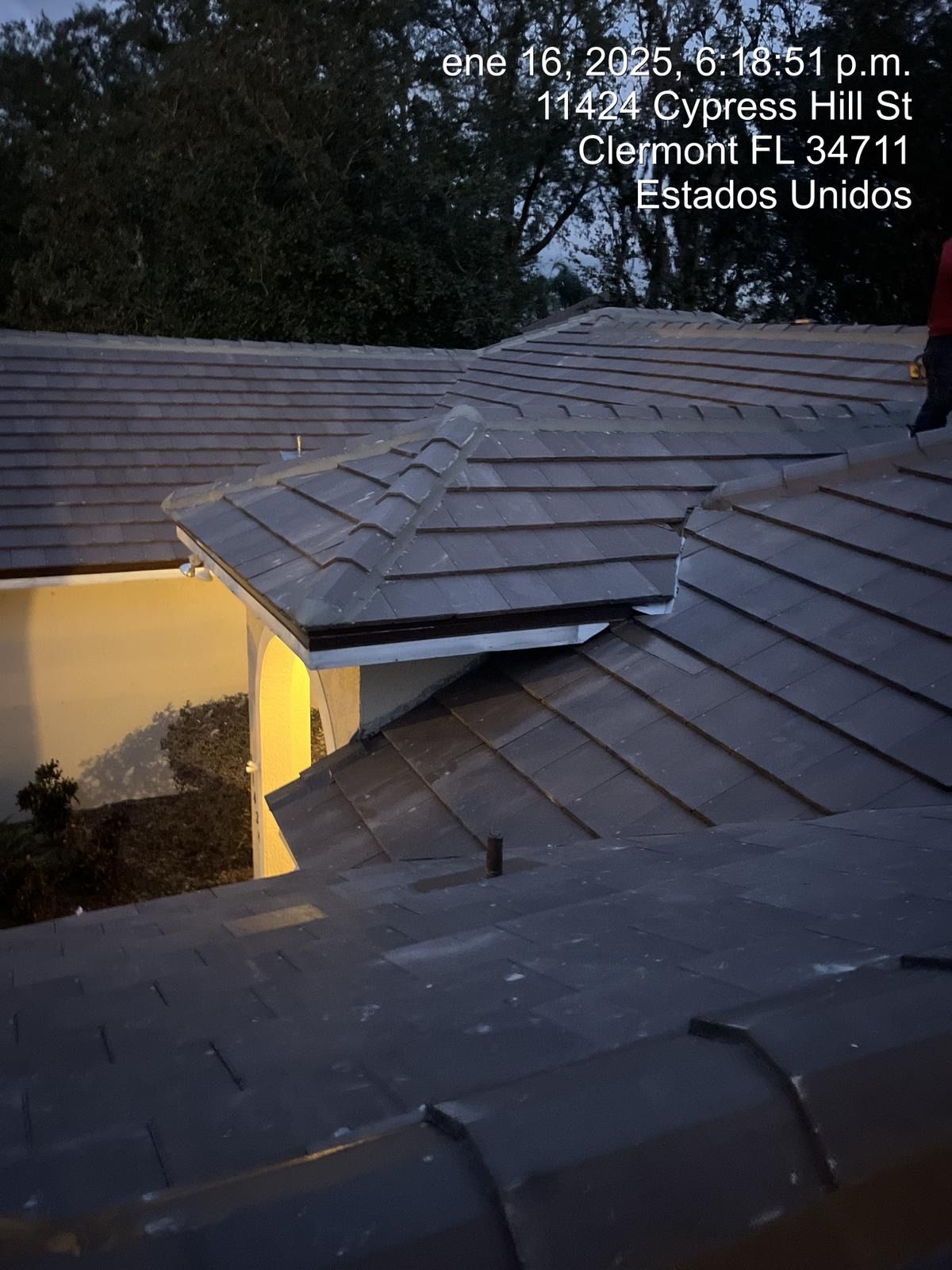 Rooftop with gray shingles at dusk, showing a small dormer and a lit yellow wall below.