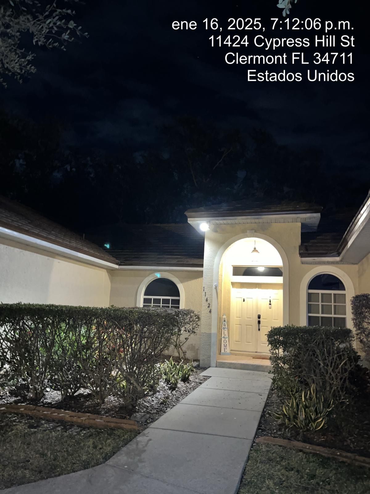 Nighttime view of a lit apartment entrance with walkway, hedges, and address text overhead.