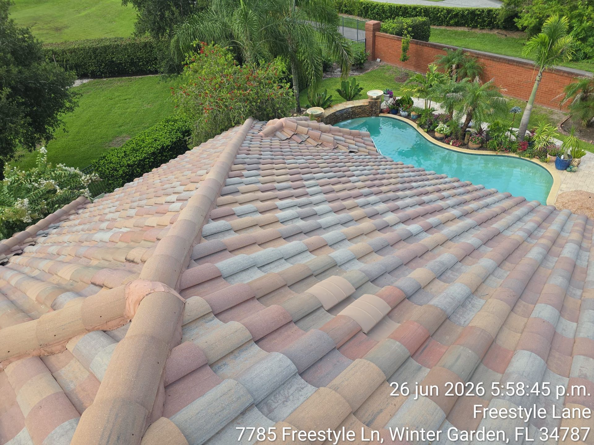 View over a terracotta-tile roof to a backyard pool, lush garden, and brick wall.