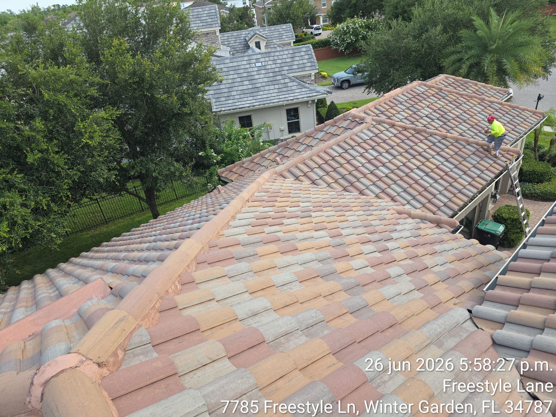 Roof being repaired with fresh shingles; worker on the right side, trees and neighboring houses in background.