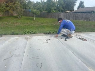 Worker in blue jacket crouches on a flat roof, repairing a seam near a grassy yard.