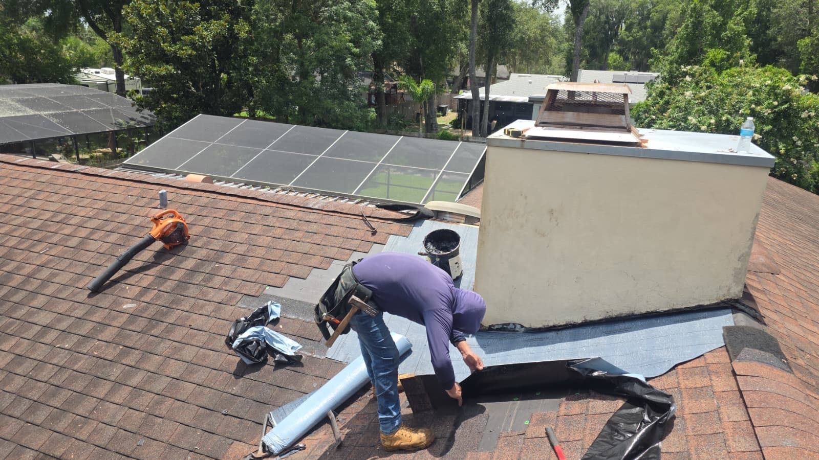 Worker installing roof flashing beside solar panels on a shingled rooftop