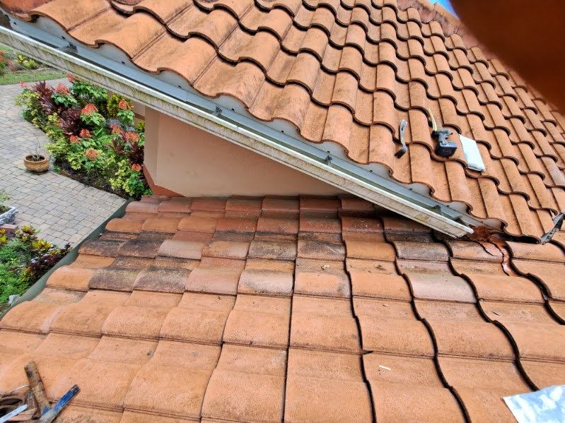Red clay tile roof with a damaged section and roof vent near flowering plants.