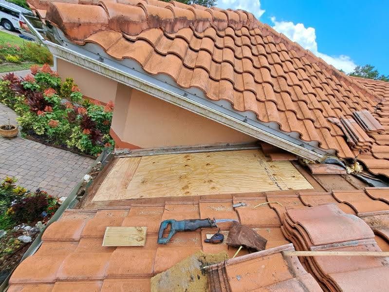 Damaged terracotta roof with exposed underlayment and tiles on a house, near a garden
