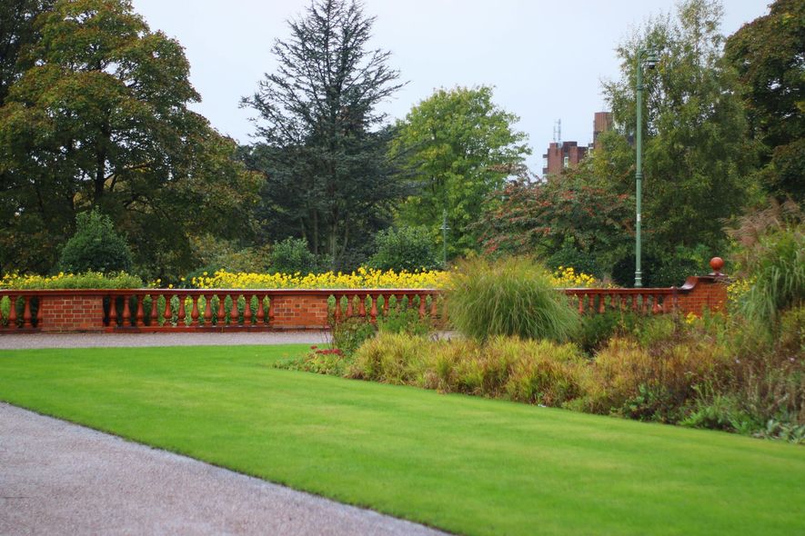 A lush green lawn leads to a decorative brick wall bordering a vibrant yellow flower bed and dense, leafy trees.
